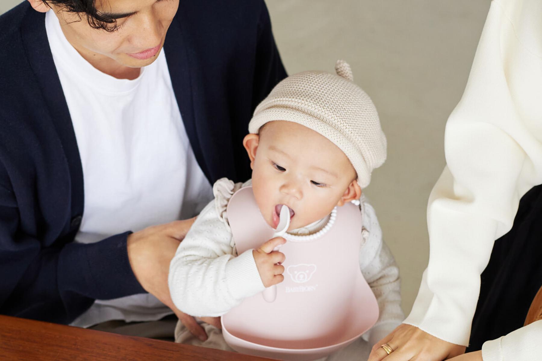 A baby wearing a pink bib and beige hat holds a spoon, sitting on an adult's lap at a table.