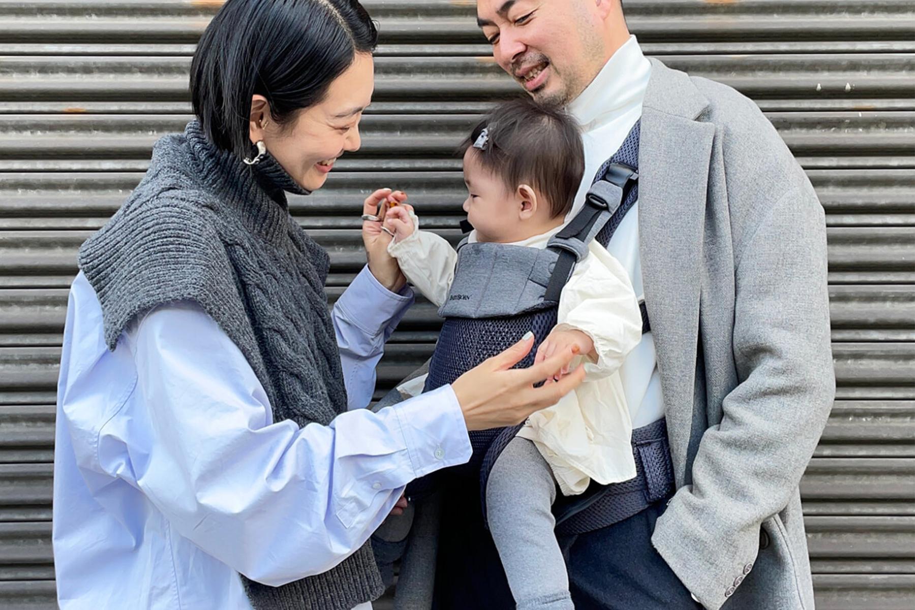 A couple smiles at their baby in a carrier. The woman wears a gray sweater, and the man wears a gray coat. They're standing in front of a metal shutter.
