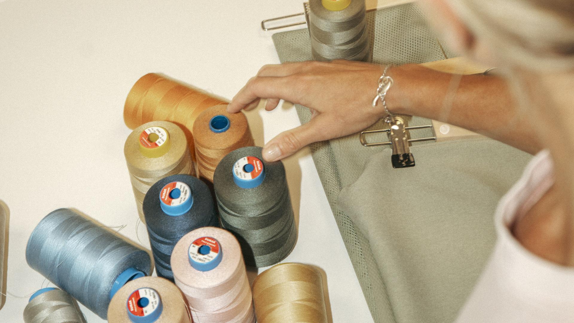 Person selecting from various spools of colorful thread on a table, next to a piece of grey fabric.