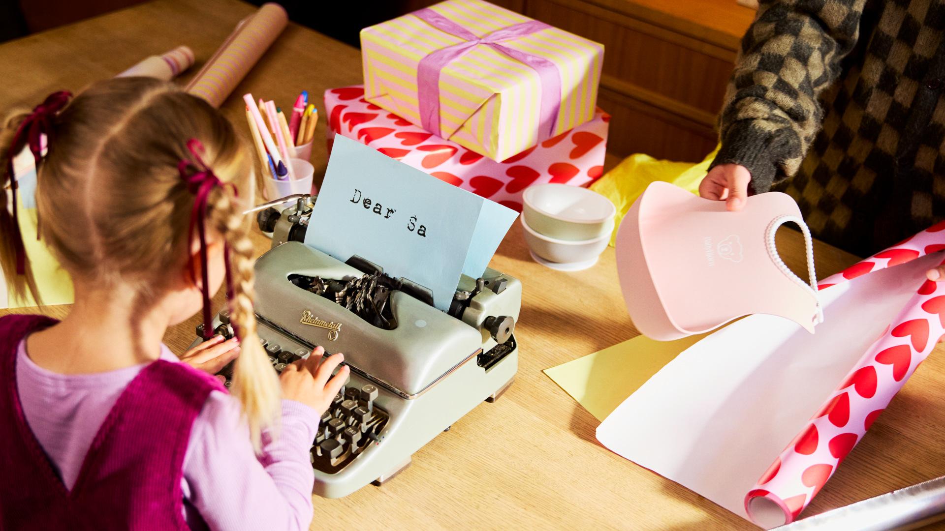 Child typing on a typewriter with a letter saying "Dear Santa." Gift wrapping supplies and a present with a purple ribbon on the table.